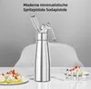 A silver whipped cream dispenser sits on a marble countertop next to two small desserts topped with whipped cream and strawberries. Several metal decorating tips and a small brush are also visible. Produktbild-Vorschau