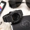 A black Casio digital watch is placed on a textured black surface, with sunglasses and a keychain in the background. Produktbild-Vorschau