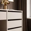 A close-up view of a white desk with drawers and a gold lamp on top, next to brown curtains. Produktbild-Vorschau