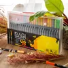A set of dual-tip marker pens in a clear plastic case, displayed on a wooden surface with decorative dried plants. Produktbild-Vorschau
