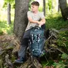 A young man sitting next to a tree in a forest with a backpack and hiking poles. Anteprima dell’immagine prodotto