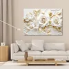 A canvas print of white and gold 3D roses is displayed on a wall above a white sofa in a living room setting. A floor lamp is to the left of the artwork, and a wooden side table with a clock is next to the sofa. Anteprima dell’immagine prodotto