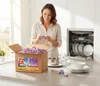 A woman is in a kitchen, unpacking a box of dishwasher tabs next to a dishwasher and clean dishes. Produktbild-Vorschau