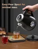A hand is pouring hot water from a black electric kettle into a glass of tea. There are other drinks and a croissant on a wooden table. Produktbild-Vorschau