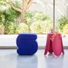 A blue stool and a pink side table with a mug and flowers are placed in a bright, airy room with a large window overlooking a garden. Produktbild-Vorschau