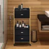 A black and gold rolling cart with drawers and shelves filled with toiletries is placed in a bathroom setting next to a sink and trash can. Produktbild-Vorschau