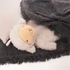 A plush sheep toy is nestled under a gray blanket, with a light-colored pillow peeking out from behind it. Podgląd zdjęcia produktu