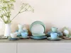 A collection of pastel-colored ceramic dishes, including plates, bowls, and cups, arranged on a countertop with a vase of greenery in the background. Náhľad obrázku produktu