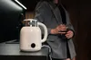 A person holding a glass of dark liquid stands in front of a white electric kettle on a dark countertop. Podgląd zdjęcia produktu