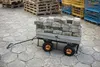 A garden cart loaded with paving stones is parked on a paved surface, with wooden pallets and metal cages in the background. Produktbild-Vorschau