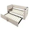 A beige upholstered daybed with a pull-out trundle bed is shown from a diagonal angle against a white background. The daybed has a built-in shelf and cup holders on one side. The trundle bed is partially pulled out. Anteprima dell’immagine prodotto