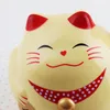 A close-up of a smiling, light yellow Maneki-neko (lucky cat) figurine with red ears and a red polka-dot collar. Produktbild-Vorschau