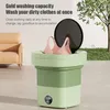 A portable washing machine with a lid open, showing two pink bras inside. The machine is green and light green. In the background, there is a beige sofa and a tree with green leaves. Náhled obrázku produktu