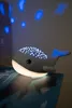 A whale-shaped night light is placed on a book, projecting stars onto the wall. Produktbild-Vorschau