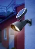 A solar-powered spotlight mounted on a blue wall, illuminating a red house exterior at dusk. Produktbild-Vorschau