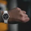 A person's wrist wearing a silver watch with a black face. Produktbild-Vorschau