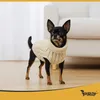 A small black and tan dog wearing a cream-colored cable-knit sweater stands on a light wooden floor in front of a white sofa and a sheer curtain. Náhľad obrázku produktu