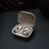 A pair of white wireless earbuds in their charging case, resting on a textured gray surface with a dark rope in the background. Náhled obrázku produktu