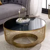 A round coffee table with a gold metal frame and a black glass top, placed in a living room setting next to a gray sofa. Produktbild-Vorschau