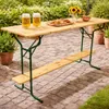 A wooden beer garden table and bench set with two glasses of beer and a plate of snacks on a paved patio with plants in the background. Produktbild-Vorschau