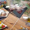 A portable charcoal grill is set up on a wooden table outdoors, with food and condiments around it. Náhled obrázku produktu