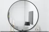 A round black-framed mirror is displayed on a white wall, reflecting a lamp and a framed picture. Two white candles in gold holders are in the foreground, and a small decorative boat is on the right. Produktbild-Vorschau
