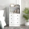 A white dresser with three drawers and an open shelf above, decorated with vases, books, and a framed picture. It is placed next to a bed and a potted plant in a room. Náhled obrázku produktu