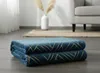A folded blue blanket with a gold geometric pattern is placed on a wooden floor in a room with a sofa and a vase in the background. Podgląd zdjęcia produktu