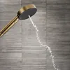 A close-up shot of a gold shower head with water spraying out, set against a textured gray tiled wall. Produktbild-Vorschau