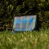 A blue plaid picnic blanket is laid out on green grass in front of a dark green bush. Produktbild-Vorschau
