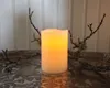 A lit LED candle is placed on a decorative tray with branches on either side, against a white paneled wall. Podgląd zdjęcia produktu