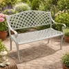 A white metal garden bench is placed on a paved patio surrounded by lush green plants and flowers. Produktbild-Vorschau