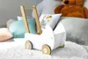 A white wooden toy pram is placed on a fluffy white rug, with pillows and a teddy bear in the background. Produktbild-Vorschau