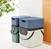 Two stacks of storage boxes with lids, one green and one white, are placed next to each other. A plant is visible in the background. Produktbild-Vorschau