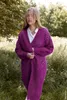 A woman stands in a grassy field wearing a long, purple knitted cardigan. Produktbild-Vorschau
