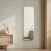 A full-length mirror with a white frame is displayed against a light beige wall, reflecting a small wooden cabinet and framed pictures. To the left, a wooden cabinet with decorative vases and a plant is visible. Voir les images du produit