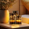 Three glass candle holders with integrated LED string lights and candles sit on a wooden surface next to a vase with greenery. Produktbild-Vorschau