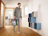 A man walks down a hallway carrying a green toolbox, with a set of cube shelves on the wall. Produktbild-Vorschau