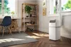 A portable air conditioner is placed in a room next to a window, with a desk and chair on the left and a shelf unit on the right. Produktbild-Vorschau