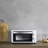 A white mini oven is placed on a wooden countertop against a gray concrete wall. A wooden cutting board is to the left, and some produce (garlic, onion) are to the right. Produktbild-Vorschau