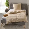 A neatly made bed with beige seersucker bedding and a pillow, placed in a bright room with a plant and a small stool. Produktbild-Vorschau