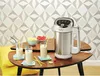 A soup maker is displayed on a wooden table with three glasses of smoothie, bowls of fruit, and yellow straws, set against a patterned wallpaper. Produktbild-Vorschau
