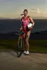 A female triathlete stands with her bicycle and helmet at sunset near a body of water. Náhled obrázku produktu