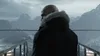 A bald man with a beard wearing a dark parka with a fur-lined hood stands on a platform overlooking a snowy mountain range. Náhľad obrázku produktu