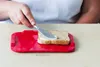 A person is buttering a slice of bread on a red cutting board. Produktbild-Vorschau