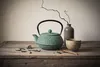 A still life arrangement of a green cast iron teapot, a ceramic cup, a wooden spoon, and dried tea leaves on a wooden table. Podgląd zdjęcia produktu