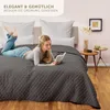 A woman is lying on a bed covered with a dark gray quilted blanket, reading a book. There are pillows behind her and a small side table with decorative items next to the bed. Náhled obrázku produktu