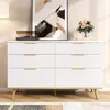 A white dresser with gold handles and wooden legs, displayed in a room with a painting and flowers. Podgląd zdjęcia produktu