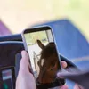 A person is holding a smartphone, showing a close-up of a horse's head on the screen. The phone is positioned in front of a steering wheel or similar object. Produktbild-Vorschau