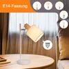 A table lamp with a white fabric shade and a wooden accent, placed on a wooden table next to some books. The background shows curtains and a sofa. Several small images of different light bulbs are displayed in the upper right corner. Produktbild-Vorschau